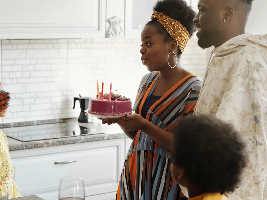 A woman proudly presenting a finished birthday cake with lit candles to her family in a home kitchen